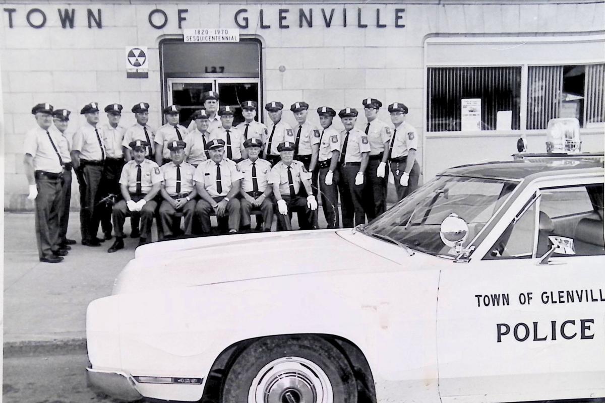 Picture of uniformed police men standing in front of a town office building with a police car in the foreground. 
